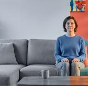 Ein geteiltes Wohnzimmer im photorealistischen Stil. In der Mitte sitzt eine Frau ruhig auf dem Sofa, exakt auf der Trennlinie zweier Welten. Die rechte Bildhälfte ist lichtdurchflutet und farbenfroh mit roten Wänden, einer grünen Couch und bunten Blumen. Die linke Bildhälfte ist komplett monochrom in matten Grau- und Blautönen gehalten, was den plötzlichen Verlust von Lebensfreude und Farbigkeit durch eine Depression symbolisiert.