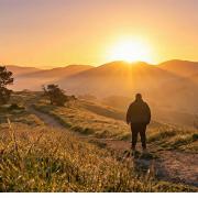 Eine kräftige Person steht von hinten gesehen auf einem unbefestigten Pfad auf einem Hügel und blickt in Richtung eines hellen, goldenen Sonnenaufgangs, der über einer weiten, hügeligen Landschaft mit nebligen Tälern aufgeht. Das hohe Gras im Vordergrund ist von warmem Licht beleuchtet.