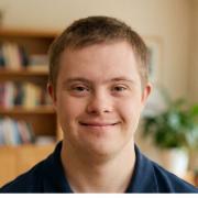 Ein professionelles, helles Porträt eines jungen Mannes mit Down-Syndrom im Alter von etwa 20 Jahren. Er blickt mit einem freundlichen, offenen Lächeln direkt in die Kamera. Er trägt ein dunkelblaues Poloshirt. Der Hintergrund zeigt ein wohnliches, helles Interieur mit einem Bücherregal und Zimmerpflanzen, das durch weiches Tageslicht angenehm unscharf gezeichnet ist, sodass der Fokus scharf auf seinem Gesicht liegt.